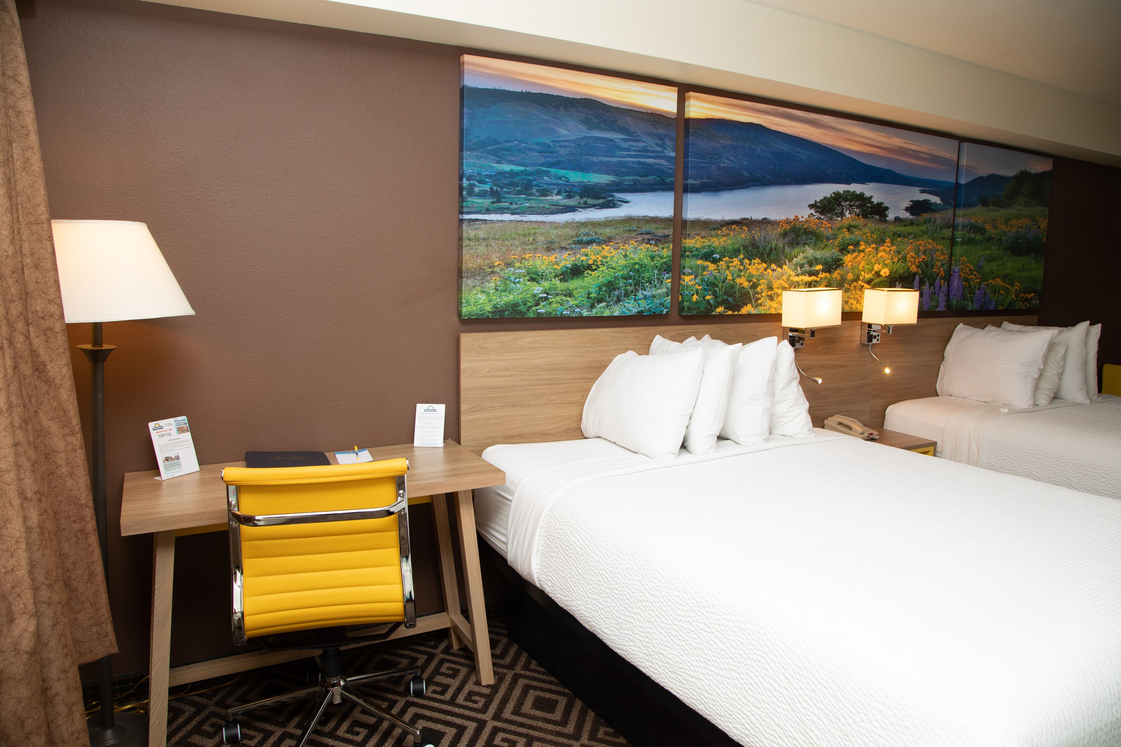 A hotel room with a bed, two bedside tables with lamps, a desk with a chair, and a large landscape painting on the wall.