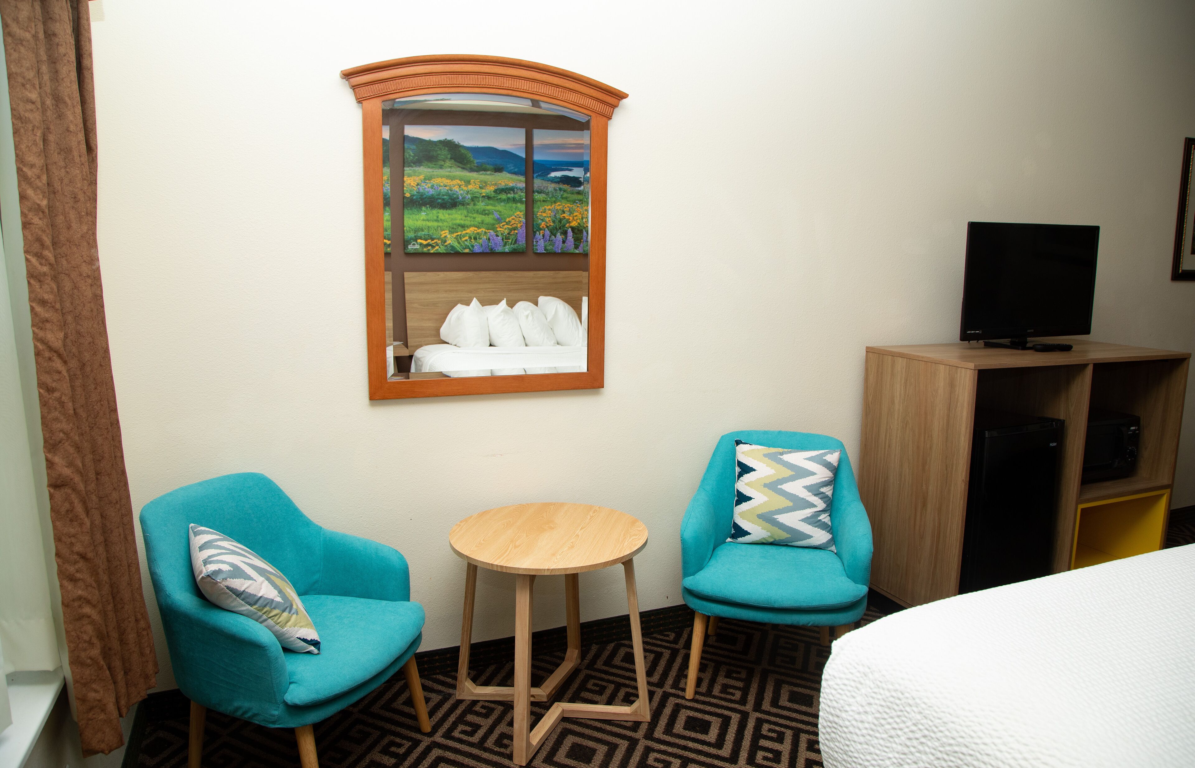 Habitación de hotel con dos sillones turquesa, una mesita de madera, una cama con ropa blanca, un televisor en un mueble de madera y una ventana con vista al paisaje.