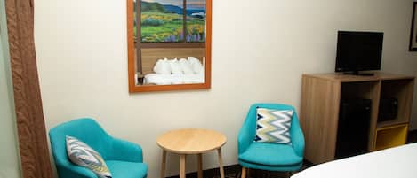 A hotel room with two turquoise armchairs, a small wooden table, a bed with white bedding, a television on a wooden cabinet, and a window with a view of a landscape.
