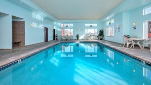 En indendørs swimmingpool med krystalklart blåt vand, omgivet af lyseblå vægge og et flisegulv. Der er stole og borde ved poolen, og en dør ind til et andet rum.
