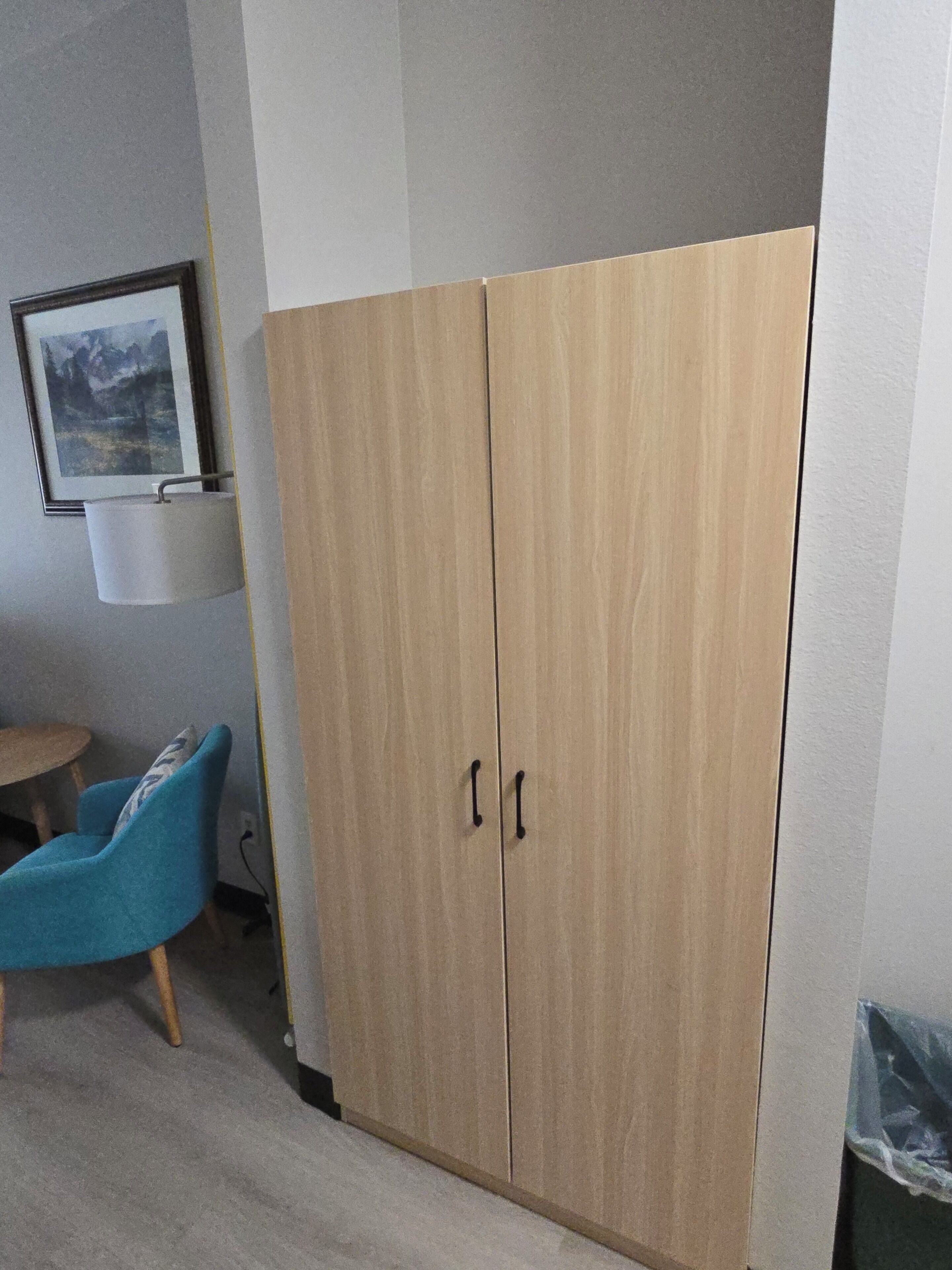 Huoneessa on vaatekaappi, tuoli, lamppu ja seinällä oleva kehystetty kuva.
