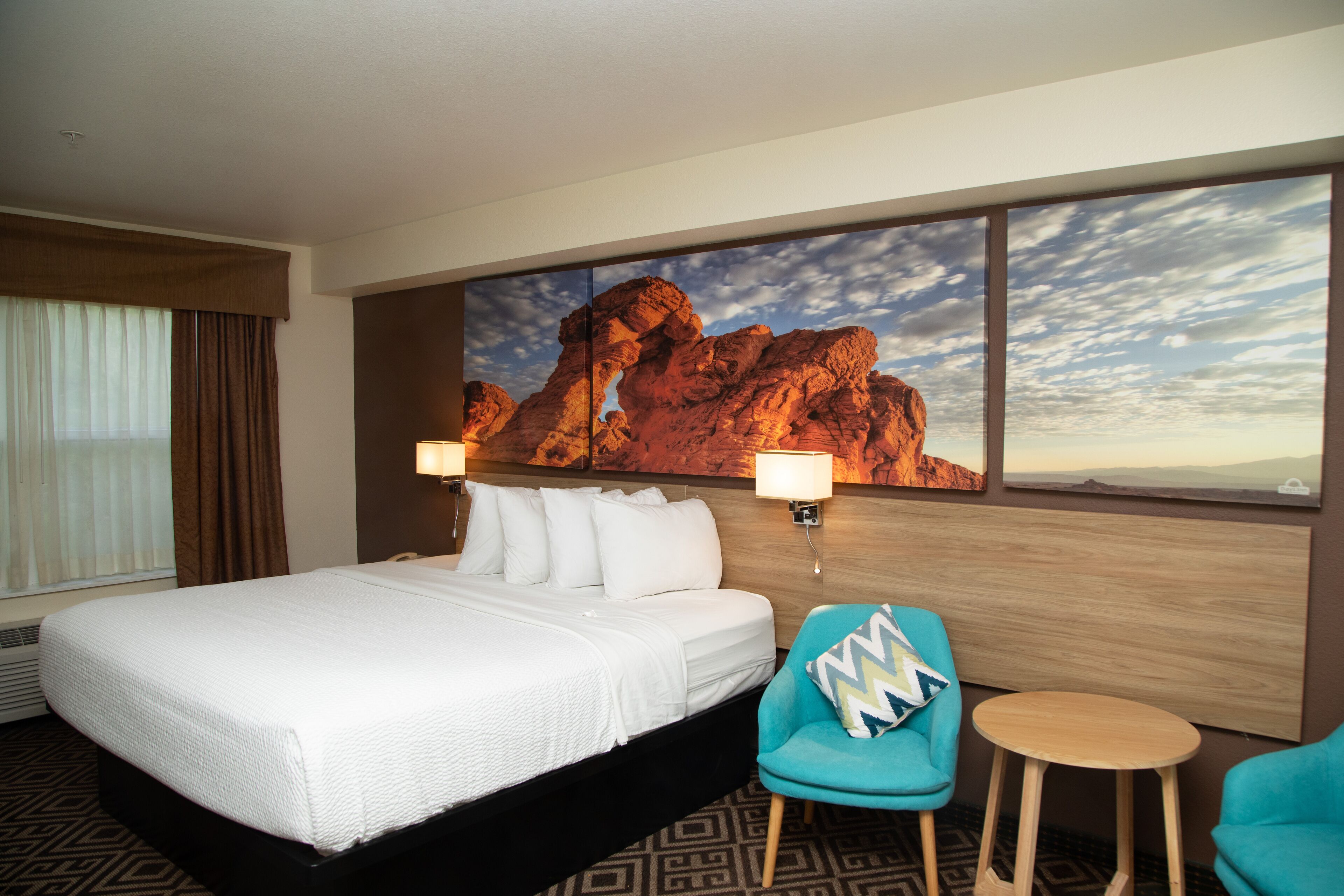 A hotel room with a large bed, a desk, two chairs, and a large rock painting on the wall.