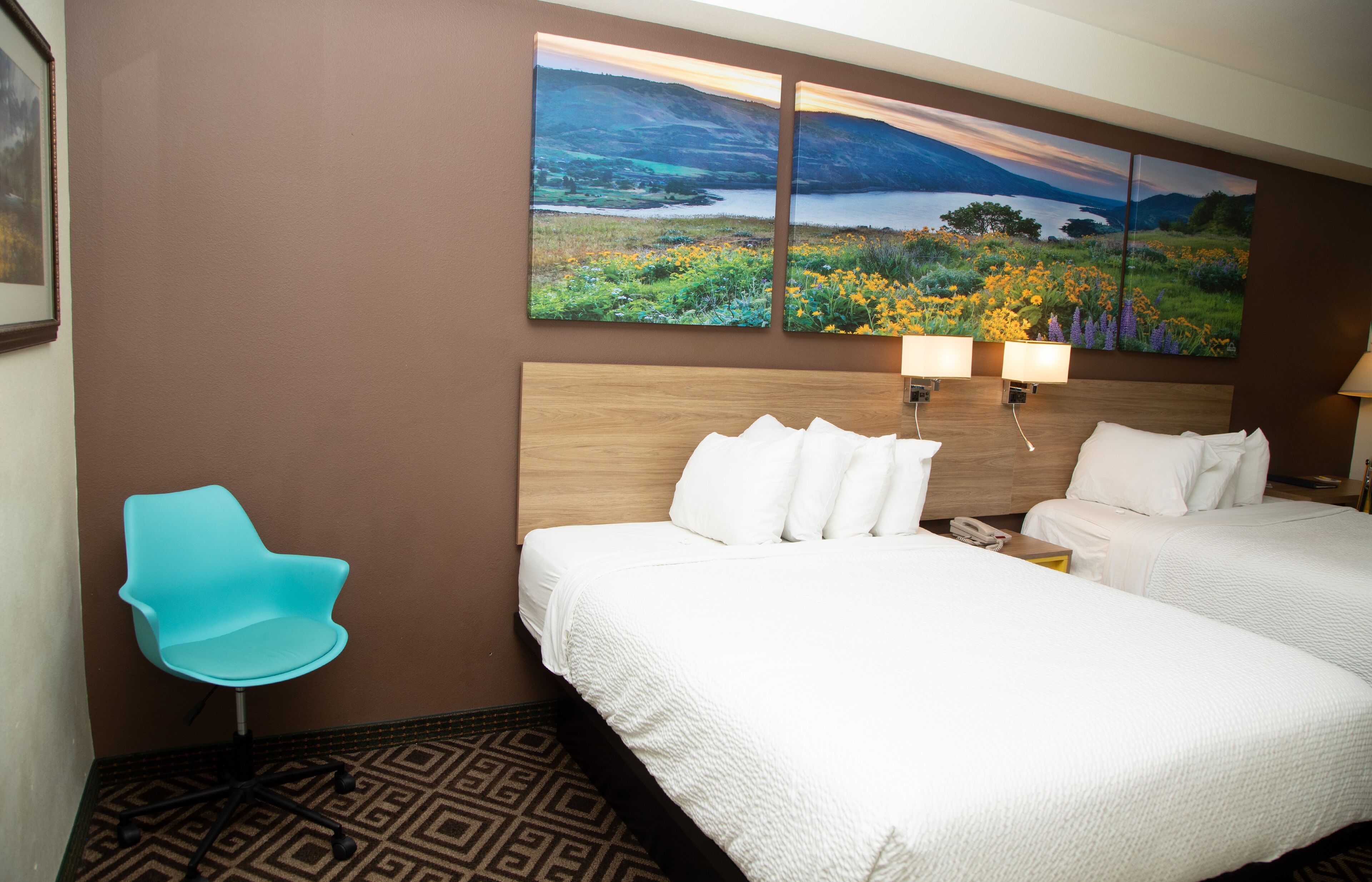 A hotel room with two beds, a blue chair, a large landscape painting, and a patterned rug.