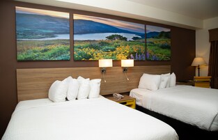 Habitación de hotel con dos camas, un cuadro grande de un paisaje en la pared y una mesita de noche con una lámpara.
