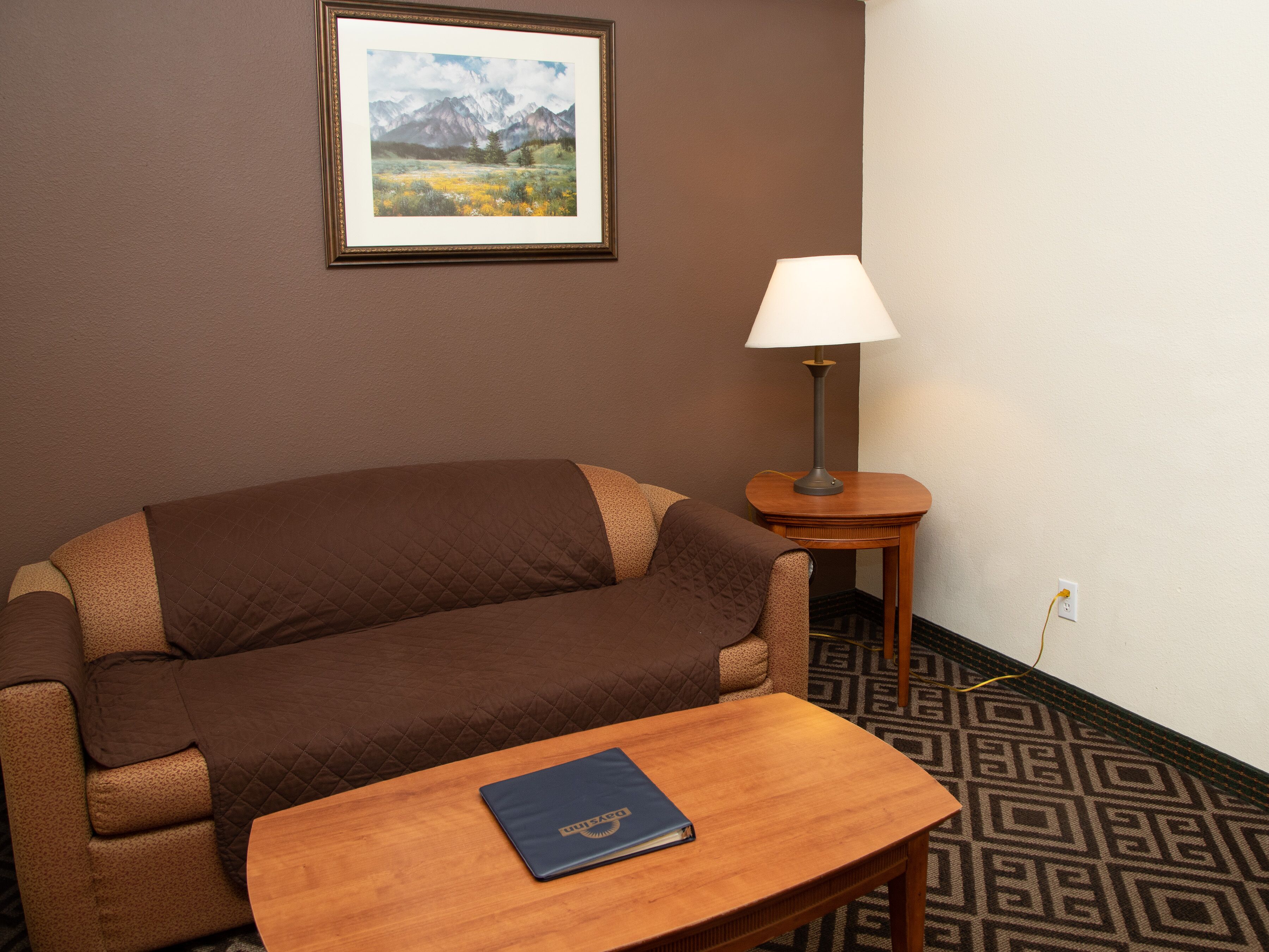 A room with a brown sofa, a small wooden table with a book, a lamp, and a framed picture on the wall.