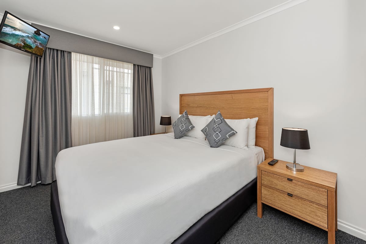 A neatly made bed with a wooden headboard, a bedside table with a lamp, and a TV mounted on the wall.