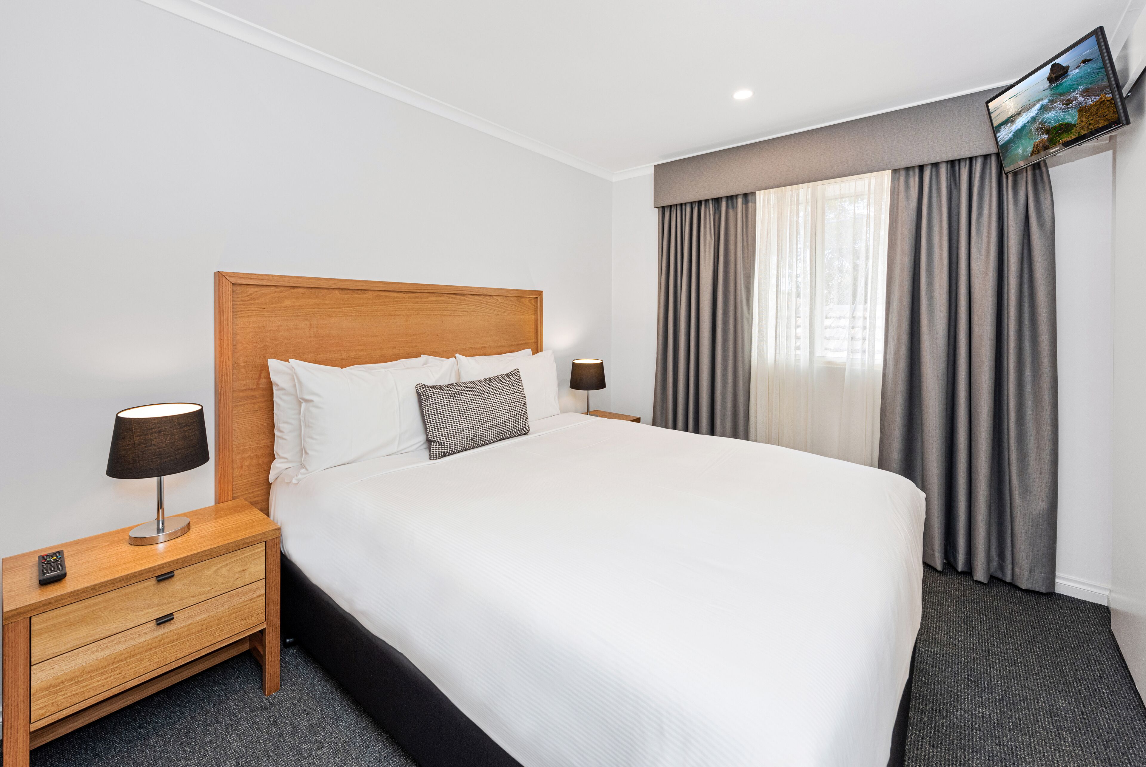 A neatly made bed with a wooden headboard, a nightstand with a lamp, and a TV mounted on the wall.