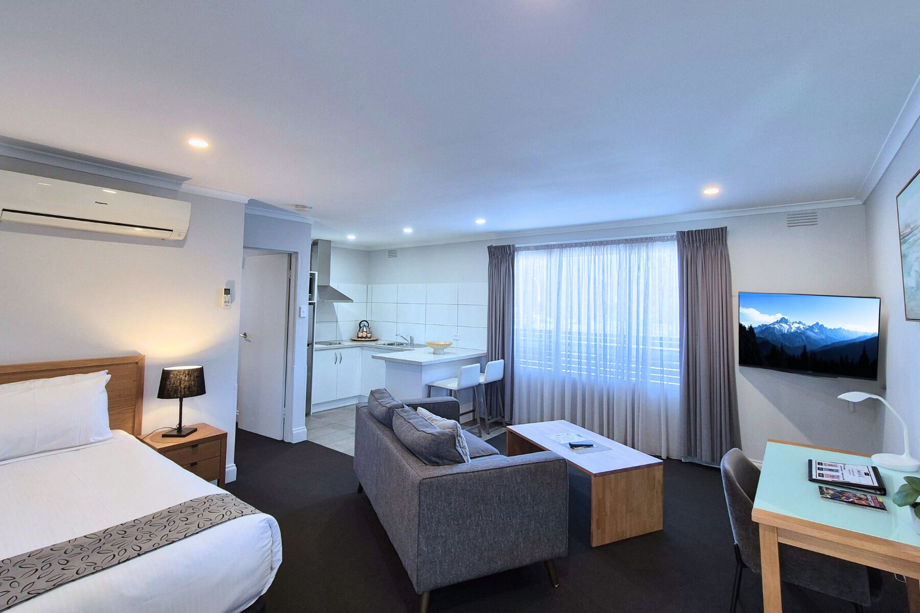 A hotel room with a bed, a sofa, a desk, a TV, and a kitchen area.