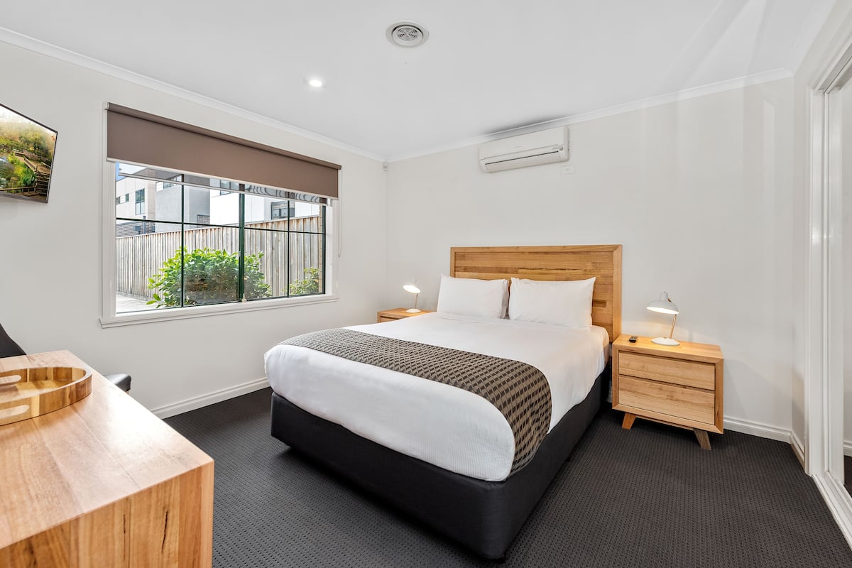 A modern hotel room with a large bed, a wooden nightstand, a desk, and a window with a view of the outside.