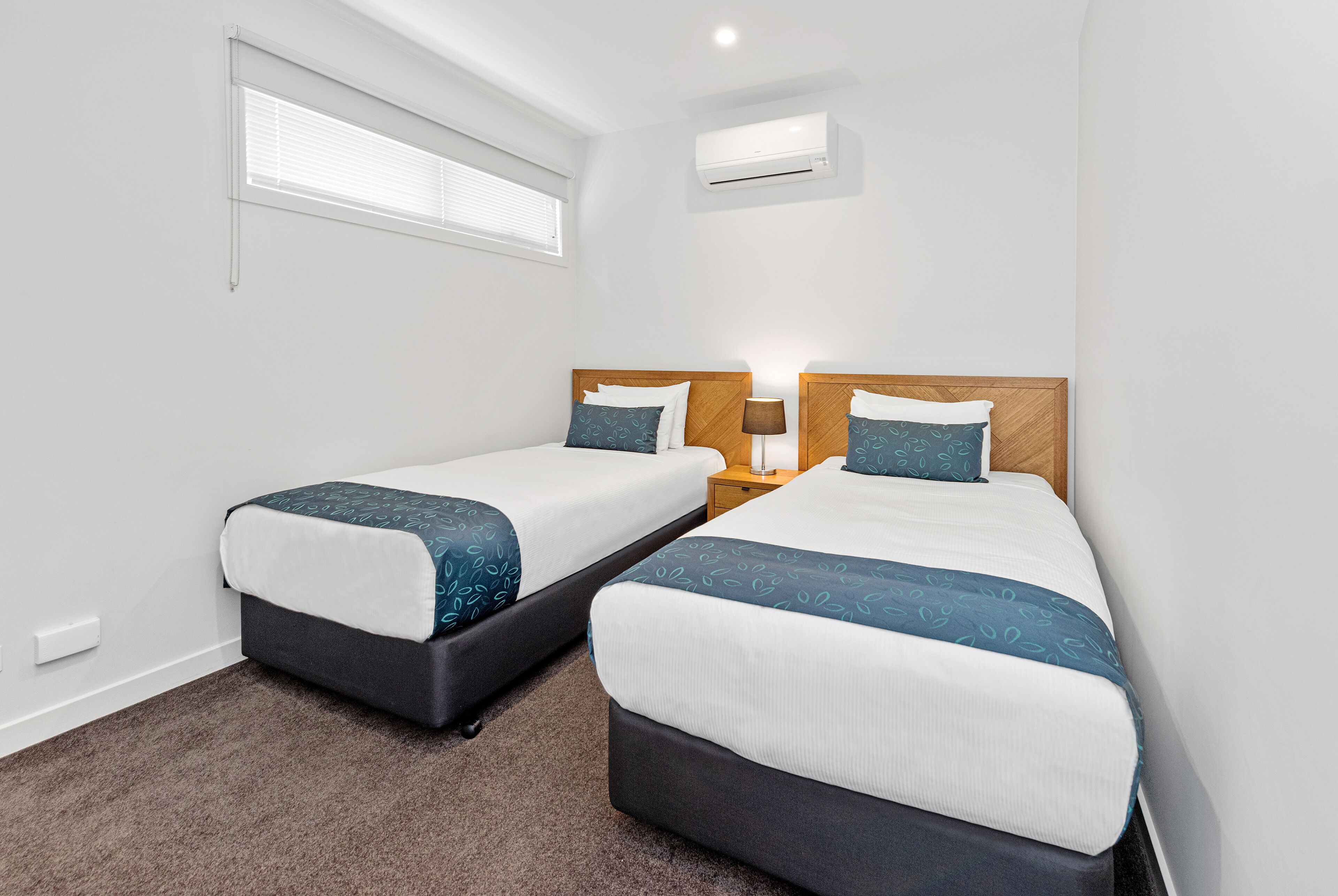 A hotel room with two beds, a window with blinds, an air conditioning unit, and a nightstand with a lamp.