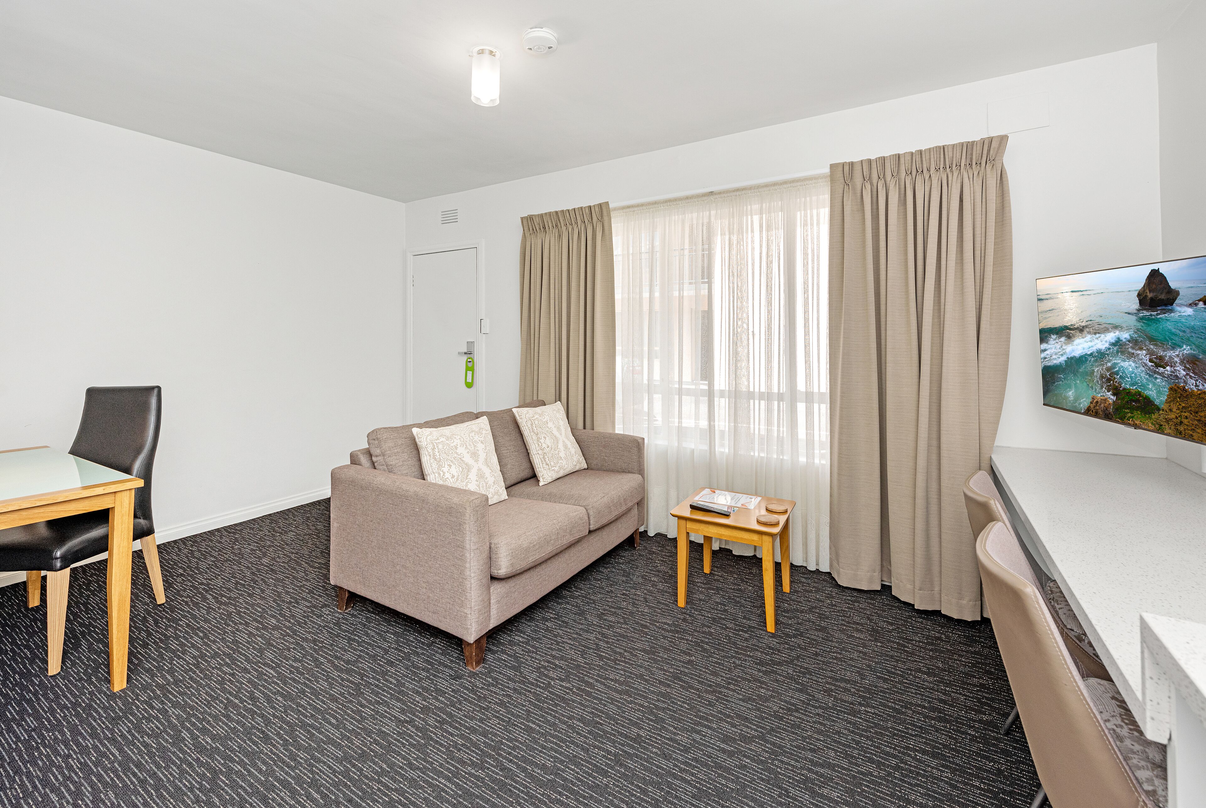 Quarto de hotel com sofá, mesa pequena, televisão e janela com cortinas.