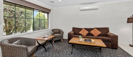 Ein modernes Wohnzimmer mit Sofa, Sessel und Couchtisch, große Fenster mit Blick auf Bäume und eine Wand-Klimaanlage.