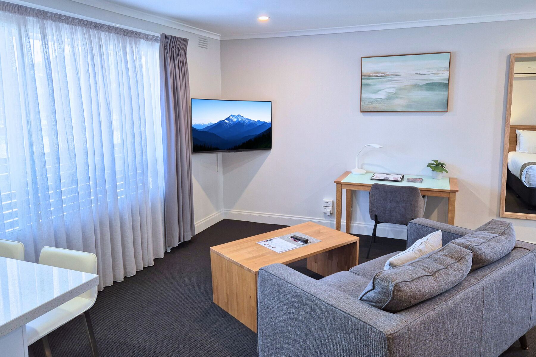 A hotel room with a wooden coffee table, grey sofa, a desk with a chair, a TV mounted on the wall, and a window with curtains.
