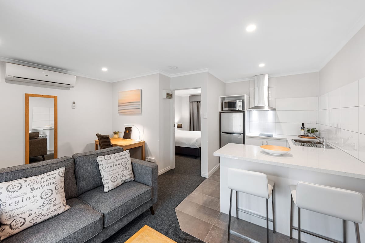A modern kitchen with a white countertop, a microwave, and a refrigerator. There is a gray sofa with patterned cushions in the living area.