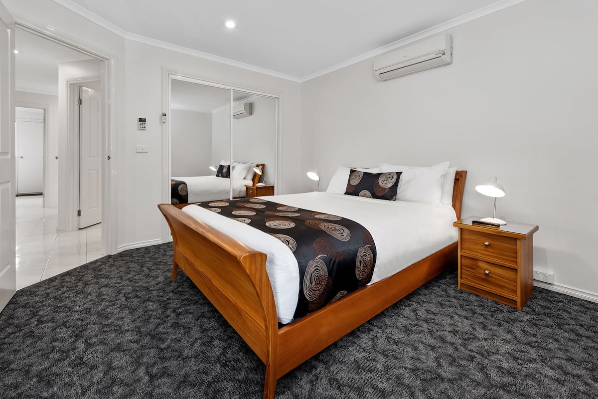 A bedroom with a wooden bed, a bedside table, a mirror, and a carpeted floor.