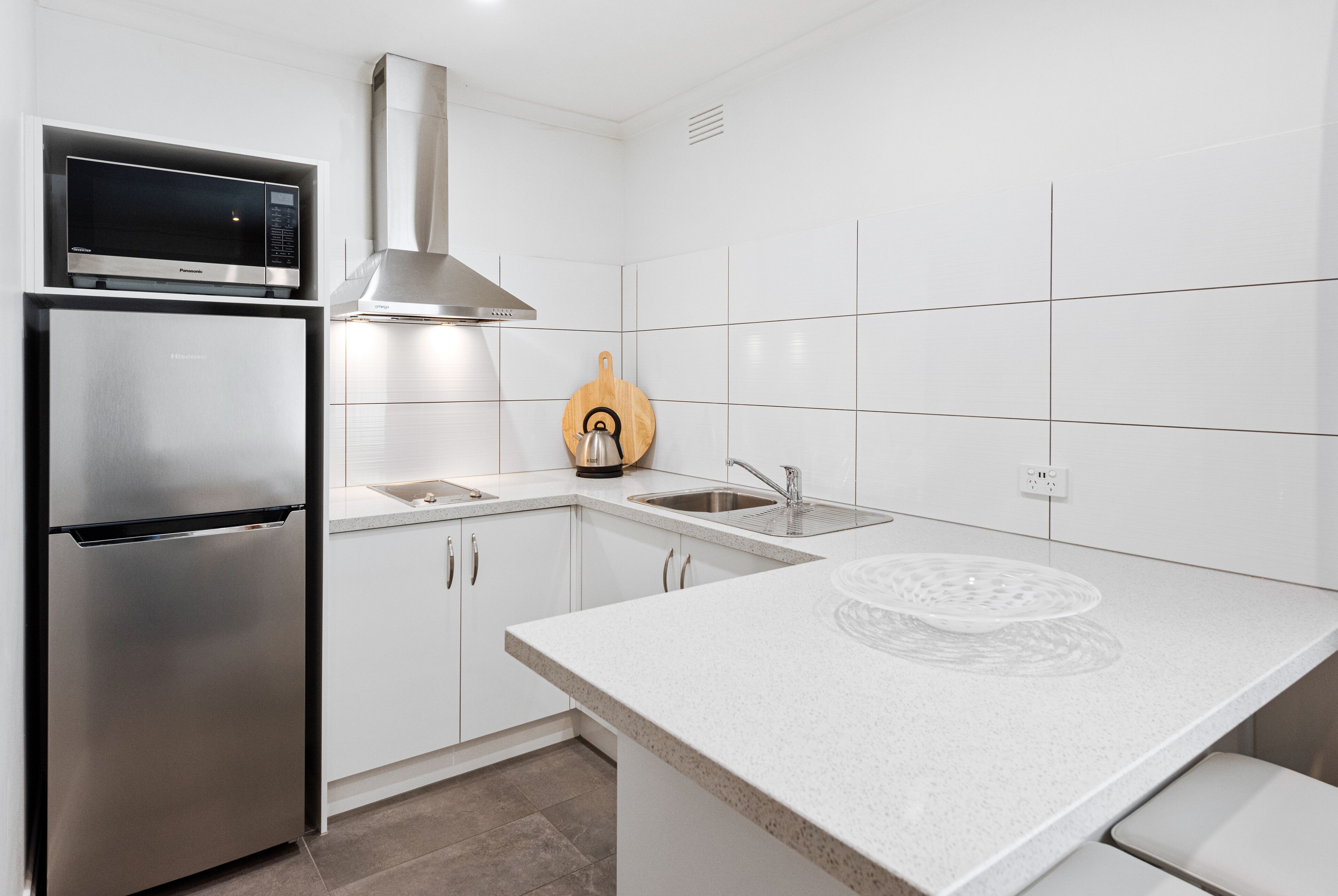 A modern kitchen with white cabinetry, a stainless steel refrigerator, a microwave, a sink, and a wooden cutting board.