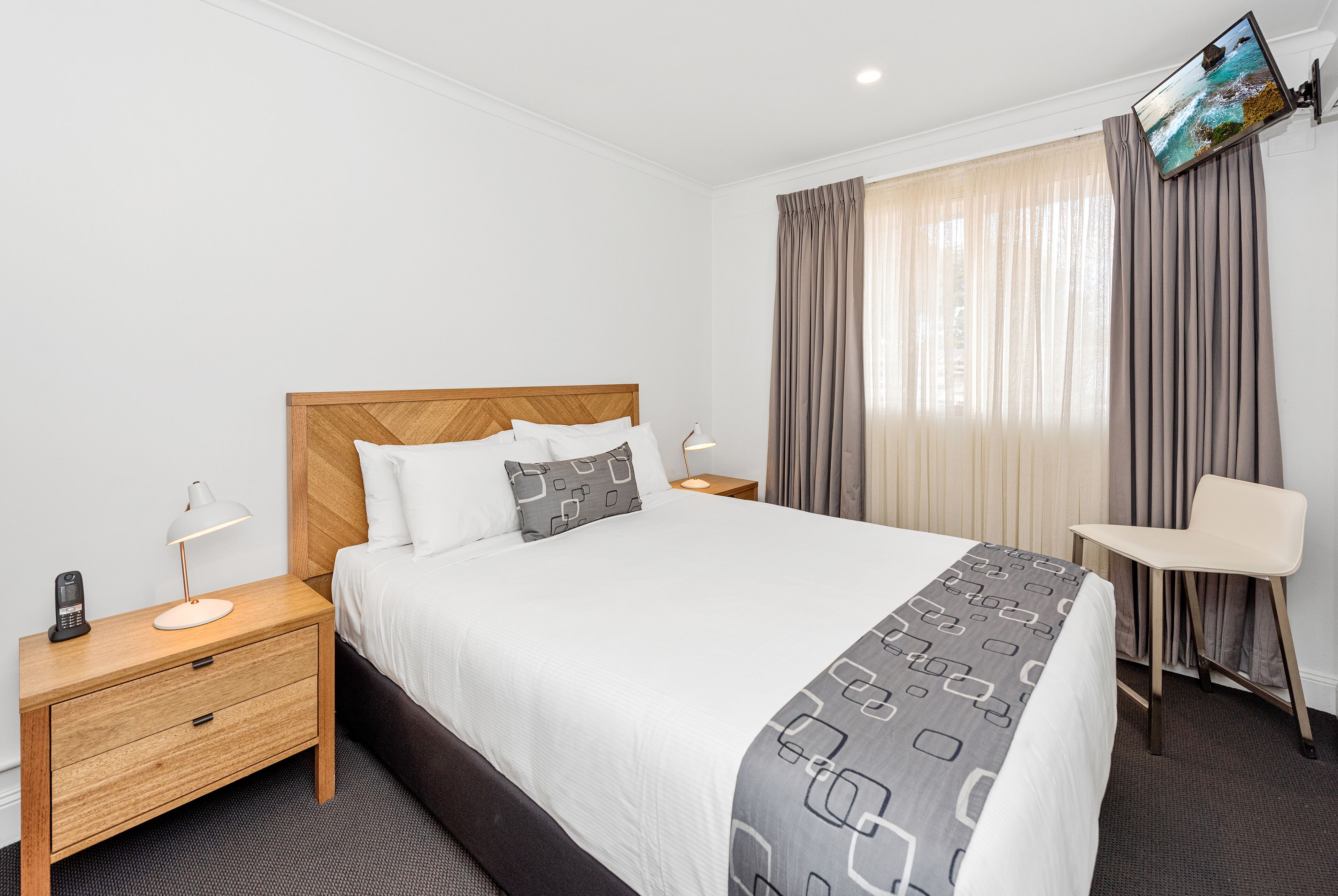 A hotel room with a large bed, a bedside table with a lamp, a chair, and a TV mounted on the wall.