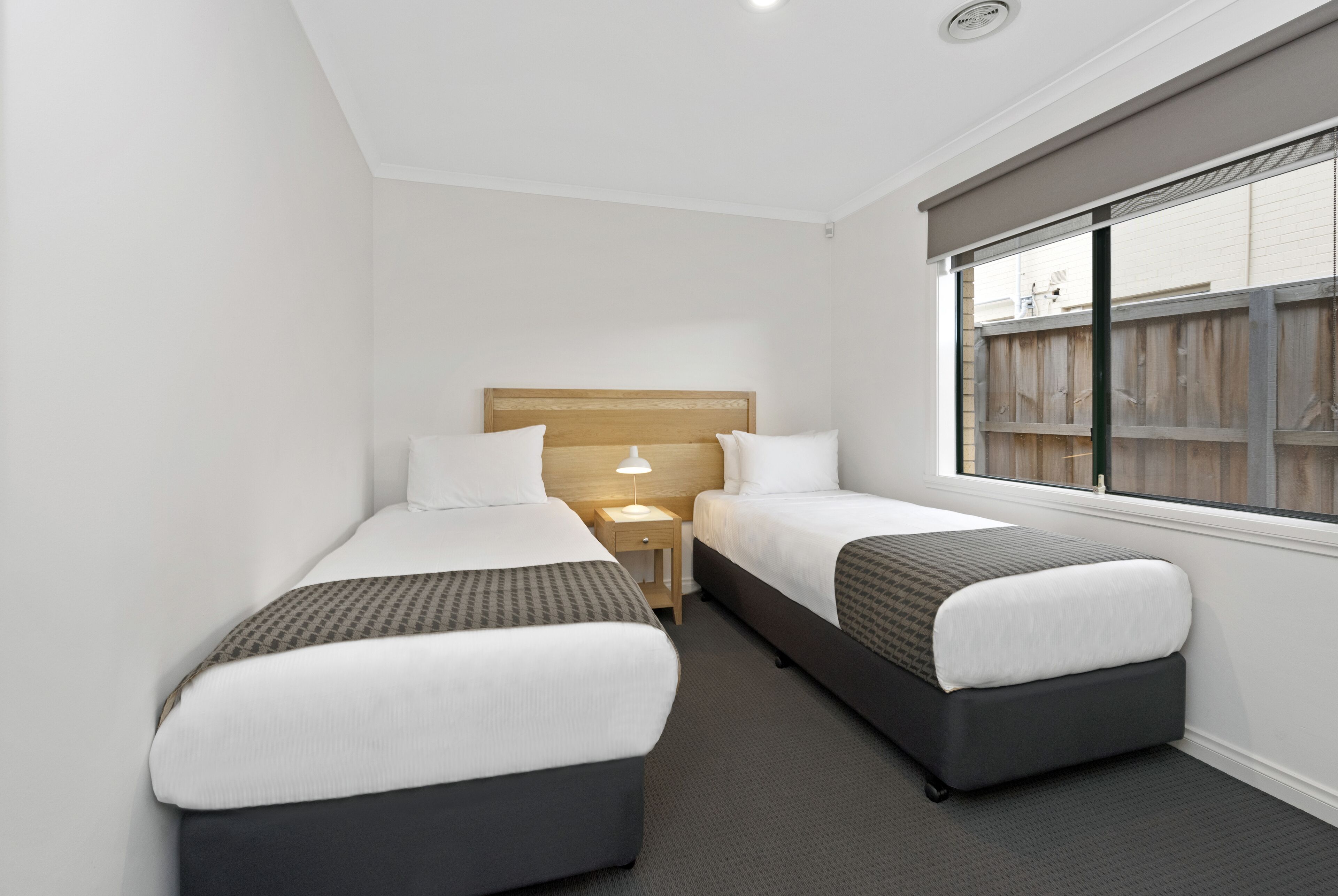 A small hotel room with two single beds, a nightstand with a lamp, and a window with a view of a wooden fence.