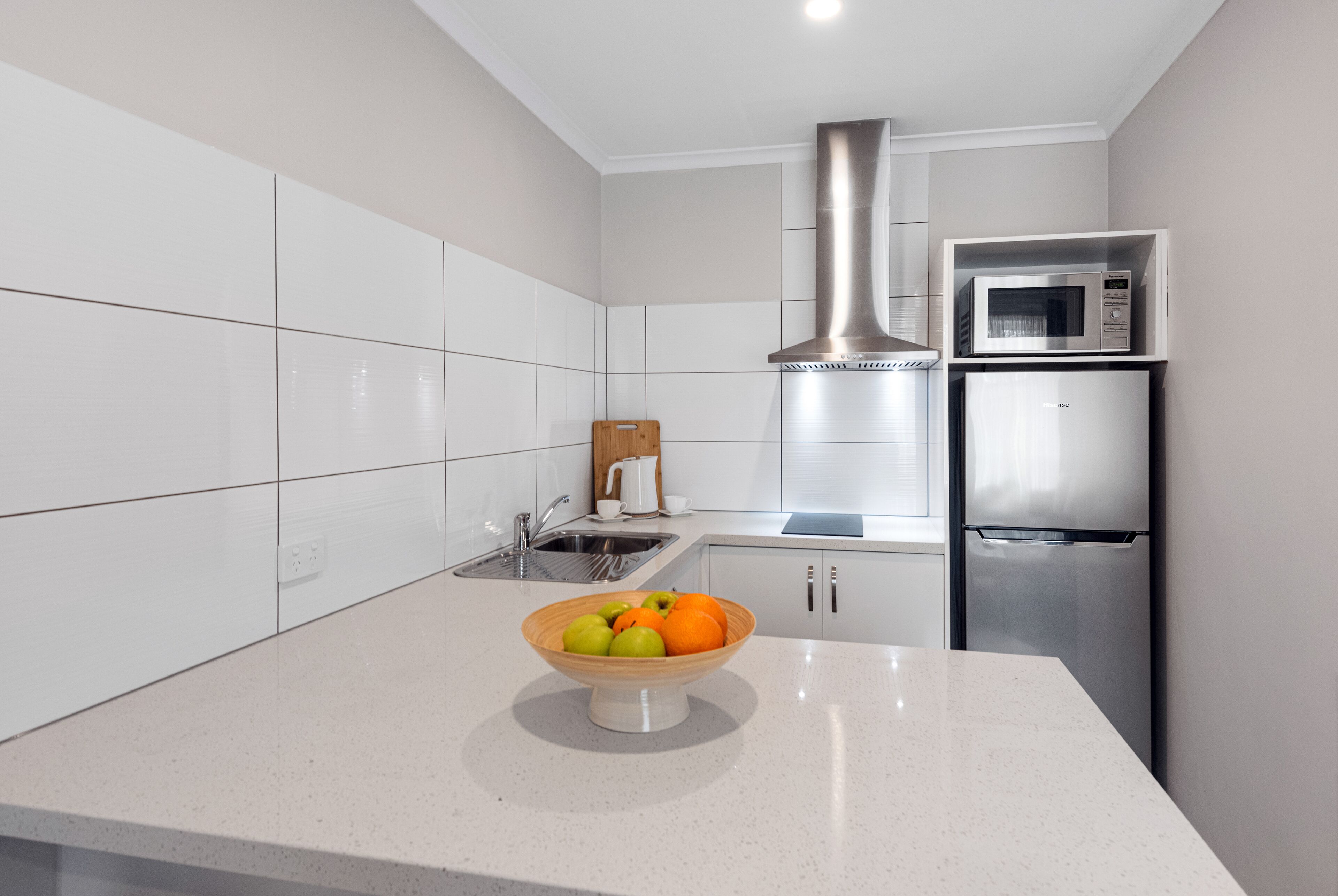 A modern kitchen with white cabinetry, a stainless steel range hood, a microwave, and a bowl of fruit on the counter.