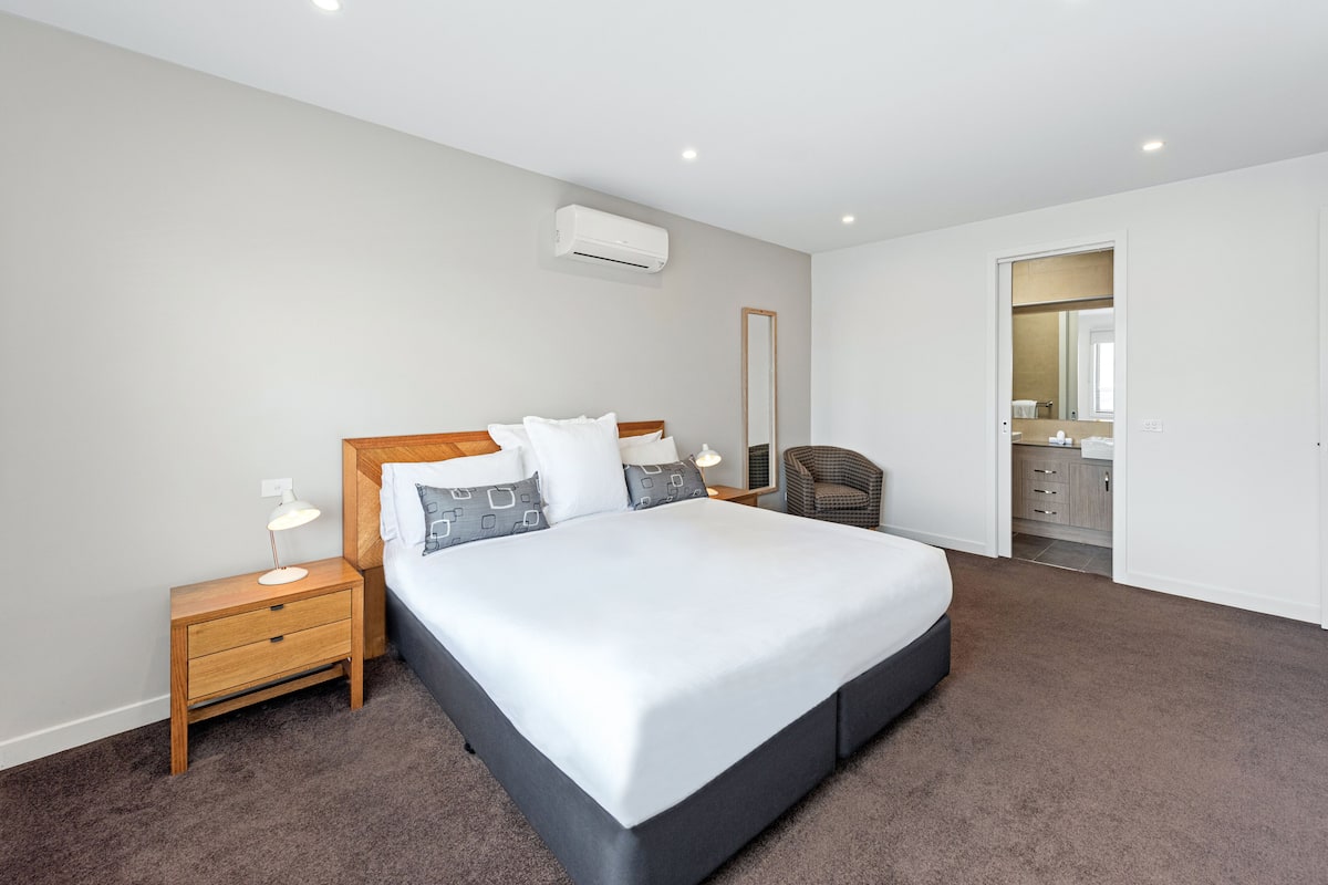 A bedroom with a large bed, a nightstand, a chair, and a bathroom visible through an open door.
