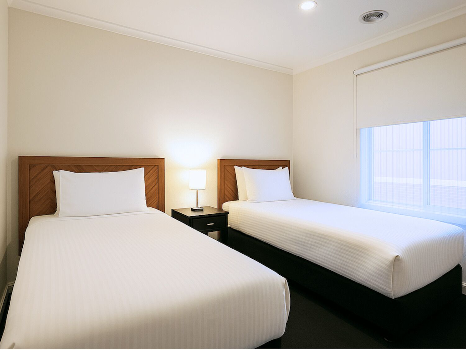 A hotel room with two single beds, a nightstand with a lamp, and a window with blinds.