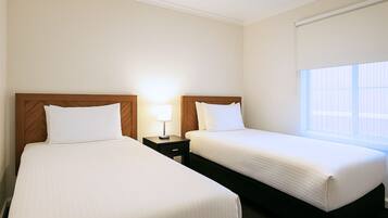 A hotel room with two single beds, a nightstand with a lamp, and a window with blinds.