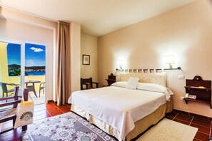 A hotel room with a bed, a desk, a chair, a nightstand, a lamp, and a view of the sea through a balcony.