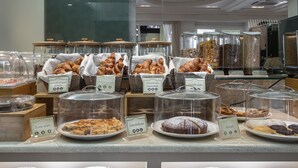 Un bufé de desayuno con diversos productos horneados y bollería exhibidos en recipientes transparentes y platos, con sus nombres identificados.