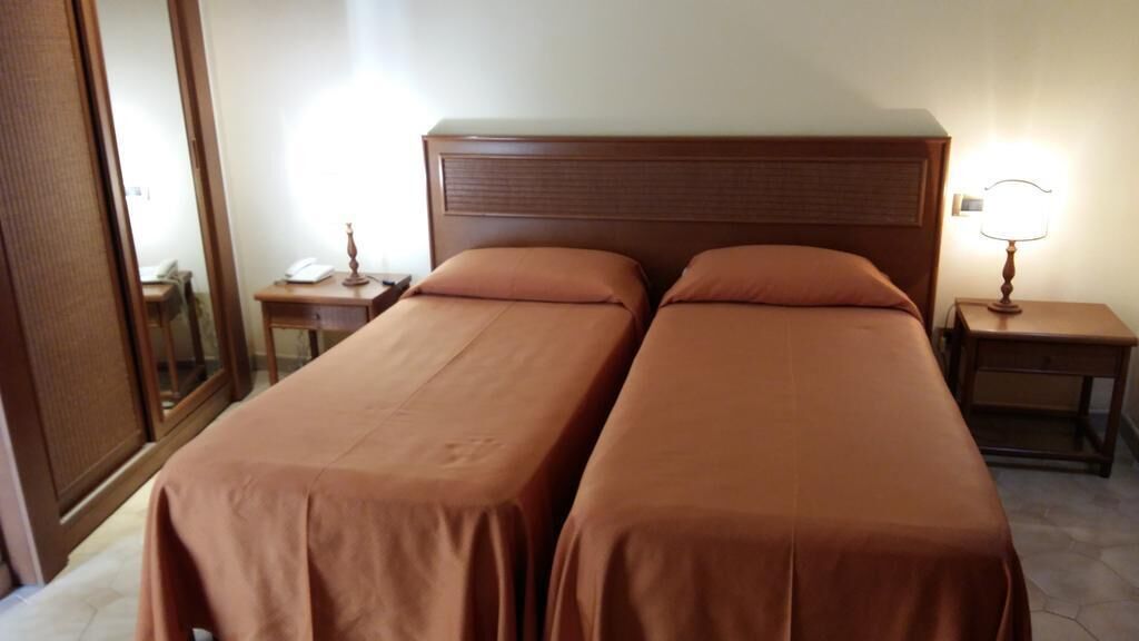 Habitación de hotel con dos camas individuales, una mesita de noche con lámpara, una pequeña mesa con teléfono y un espejo en la pared izquierda.