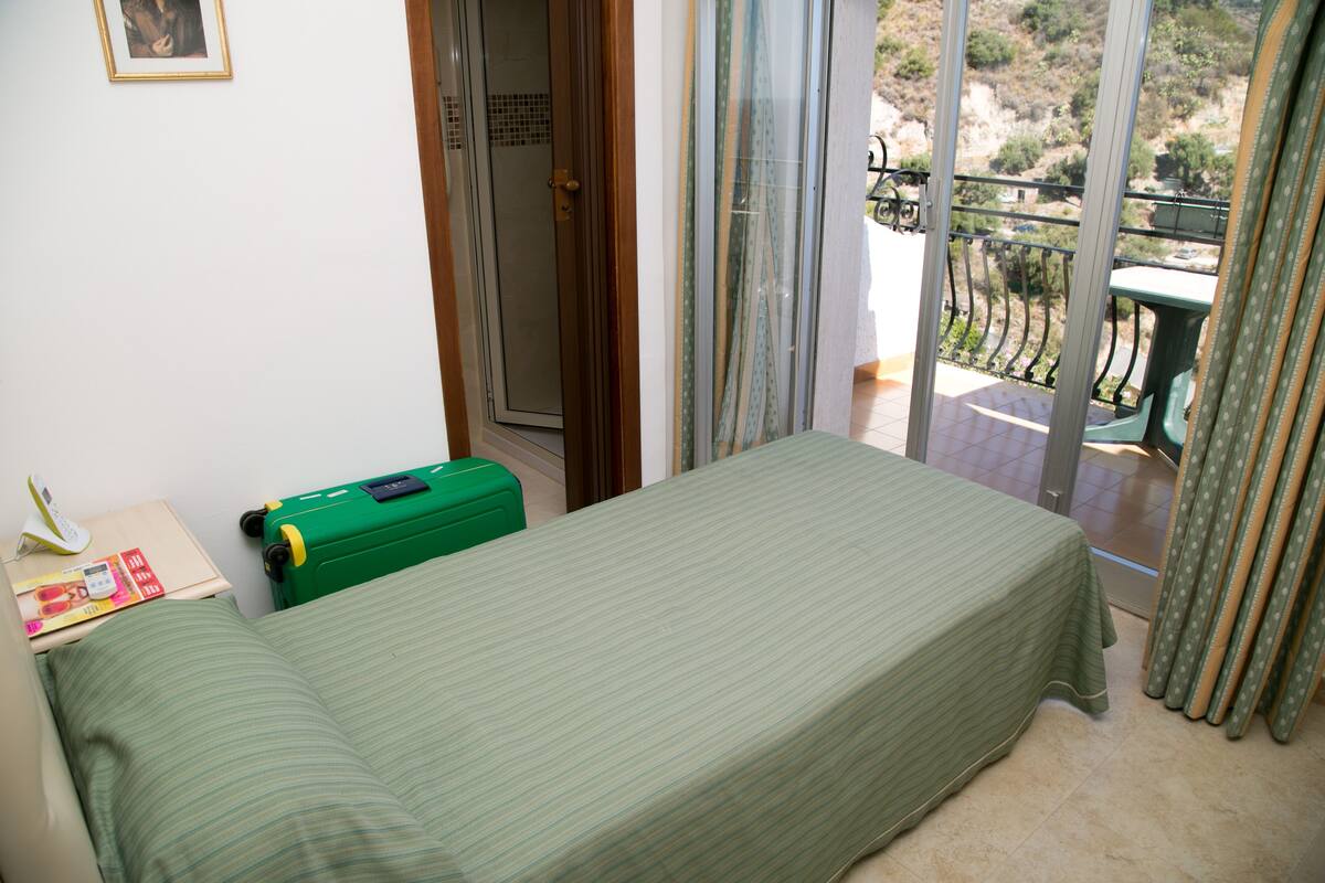A small room with a bed, a suitcase, a table with a phone and a magazine, and a balcony with a view of the countryside.