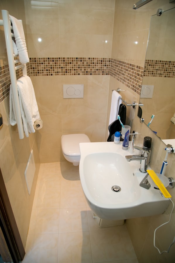 A bathroom with a white sink, toilet, and shower. There are towels hanging on a rack, a toothbrush holder, and a soap dispenser. - Hotel Villa Bianca (Taormina)