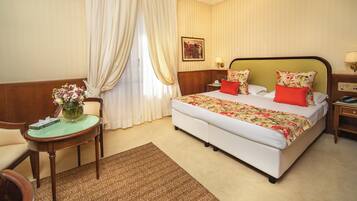 Una habitación de hotel con una cama grande, una mesita con un jarrón de flores, una silla y una mesita de noche con una lámpara.