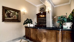 A reception area with a wooden counter, a framed painting, a potted plant, a wine rack, and a large vase. - Grand Hotel Bastiani (Grosseto)