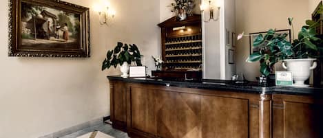 A reception area with a wooden counter, a framed painting, a potted plant, a wine rack, and a large vase.