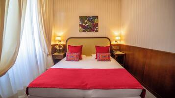 Una habitación de hotel con una cama grande, dos lámparas de noche, un cuadro floral en la pared y una manta roja.