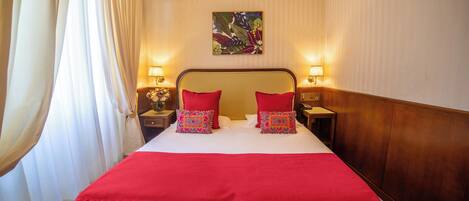 Una habitación de hotel con una cama grande, dos lámparas de noche, un cuadro floral en la pared y una manta roja.