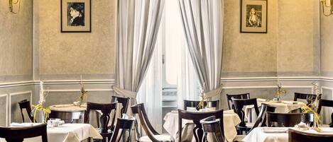 A dining area with tables set for service, framed artwork on the walls, and a large window with curtains.