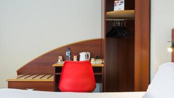 A hotel room with a wooden headboard, a red chair, a small desk with a kettle, a phone, and a coat rack with hangers.