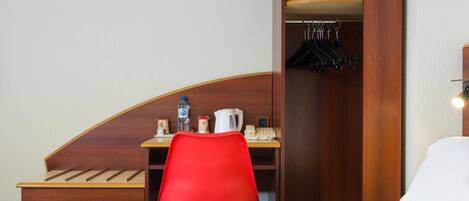 A hotel room with a wooden headboard, a red chair, a small desk with a kettle, a phone, and a coat rack with hangers.