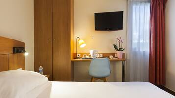 A hotel room with a bed, a desk with a lamp, a TV, and a window with curtains.