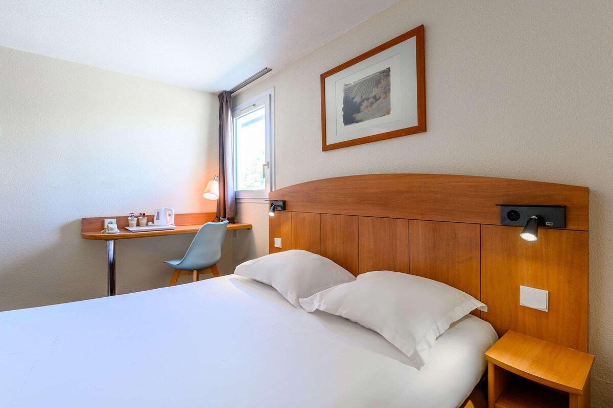 Habitación de hotel con cama, mesita de noche, silla, escritorio, lámpara y un cuadro en la pared.