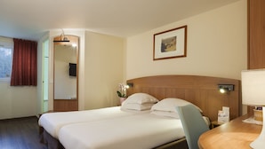 Een hotelkamer met twee bedden, een houten hoofdbord, een klein tafeltje met een lamp, een lijst met een afbeelding aan de muur, en een raam met gordijnen.
