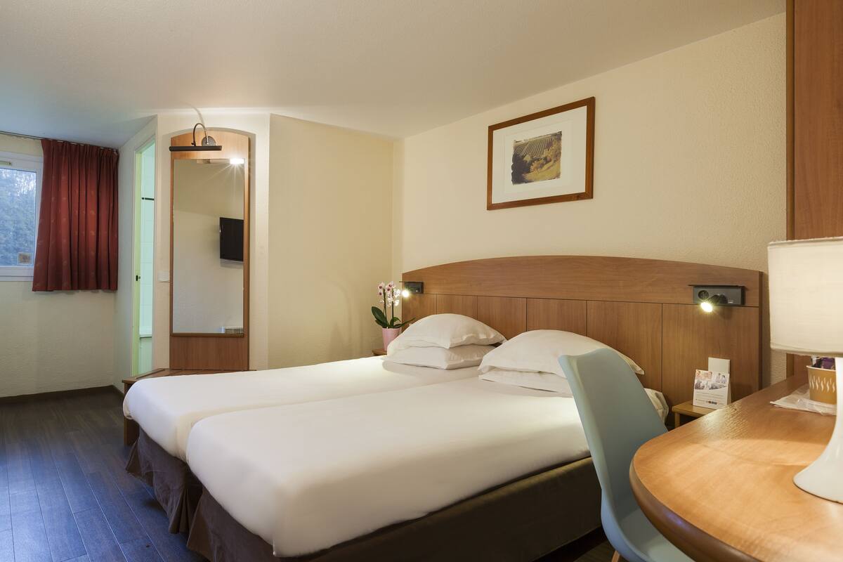 Una habitació d'hotel amb dos llits, un panell de capçalera de fusta, una taula petita amb una làmpada, un quadre amb marc a la paret i una finestra amb cortines.