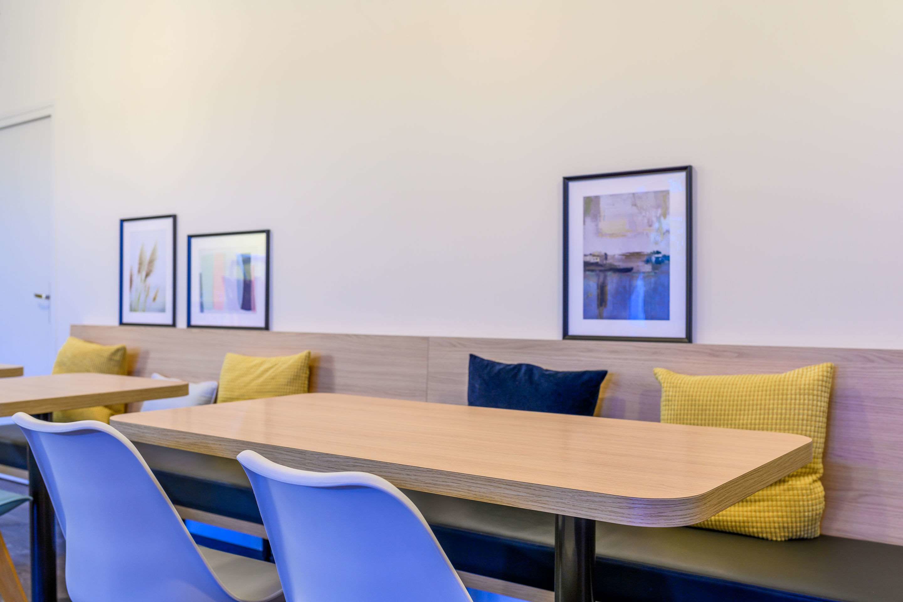 Un'area ristorazione moderna con tavoli in legno e sedie blu, che include una panca con cuscini gialli e blu.