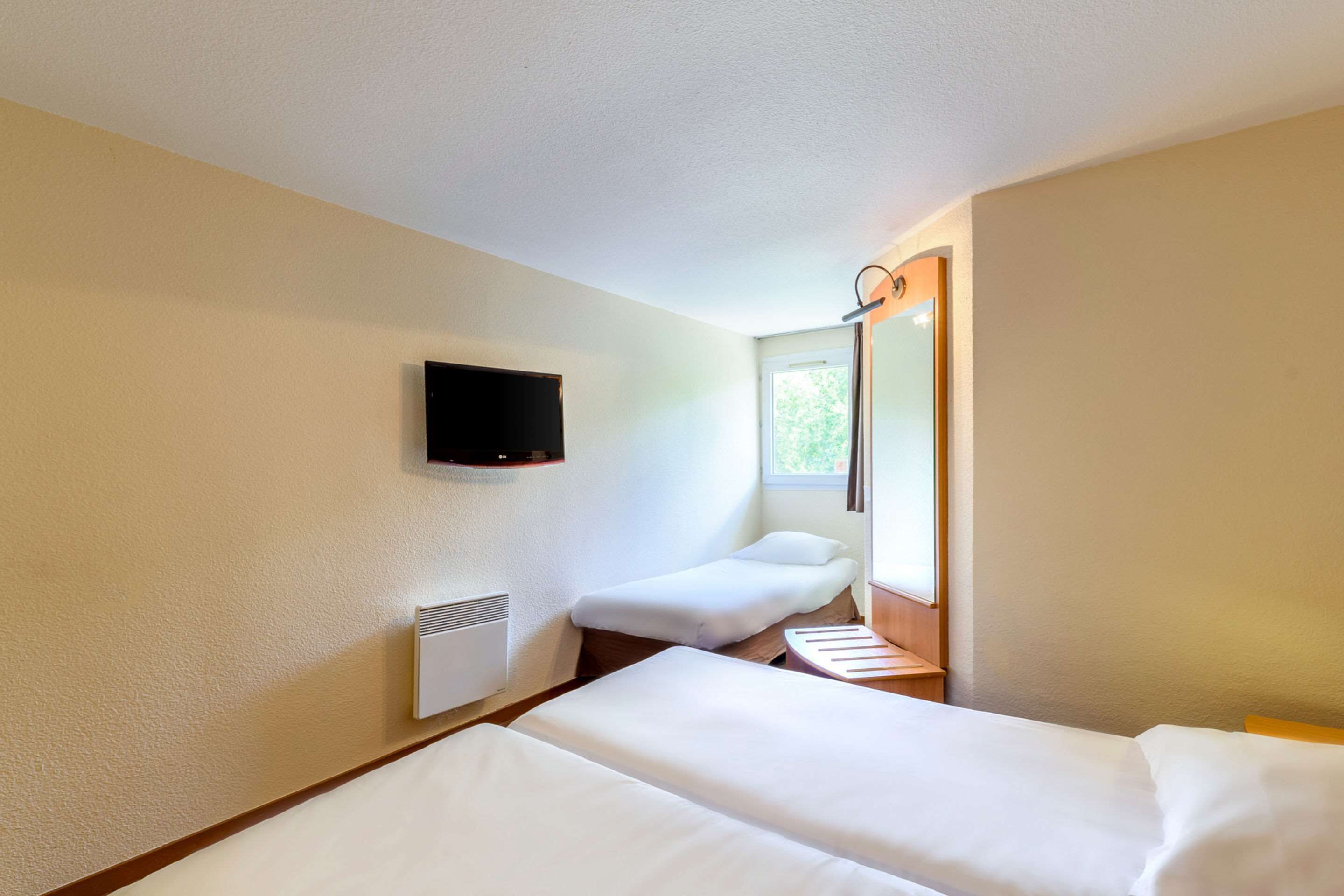 A hotel room with two beds, a TV mounted on the wall, a window, and a heater.