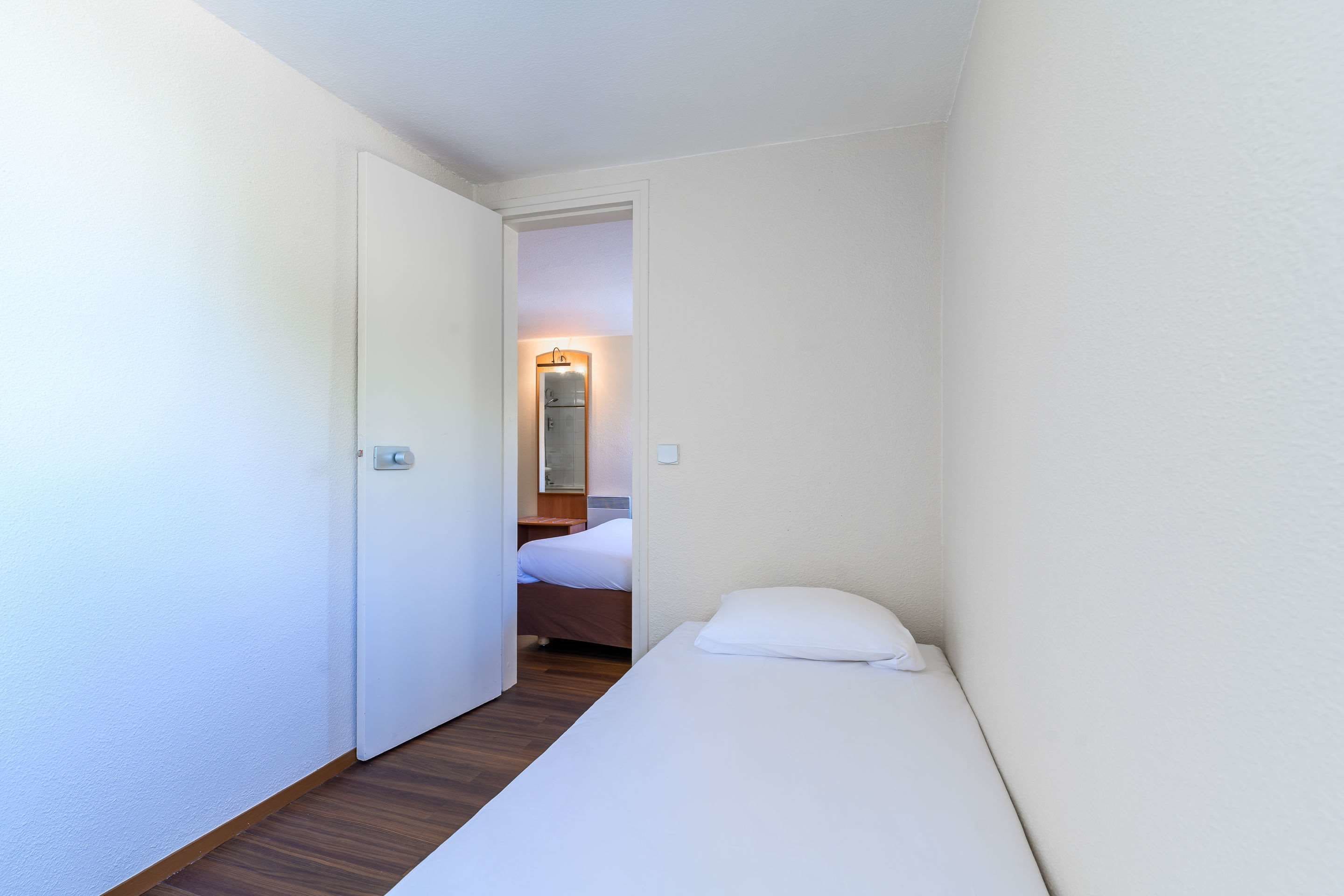 A narrow room with a single bed, a wooden floor, and a white door leading to a bathroom with a mirror and light fixture.
