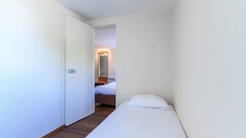 Una habitación pequeña con una cama individual, suelo de madera y una puerta blanca que da a un baño con espejo e iluminación.