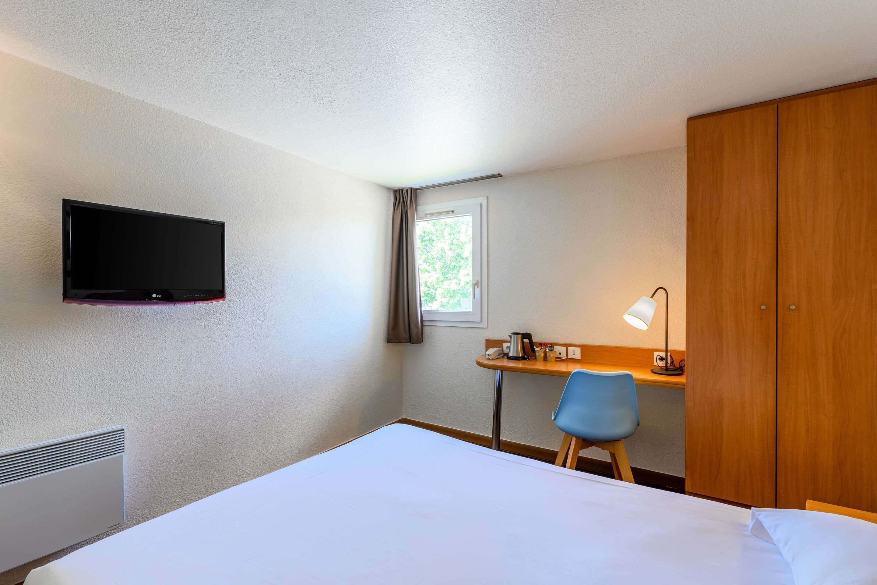 A hotel room with a bed, a desk with a chair, a TV mounted on the wall, and a wardrobe.