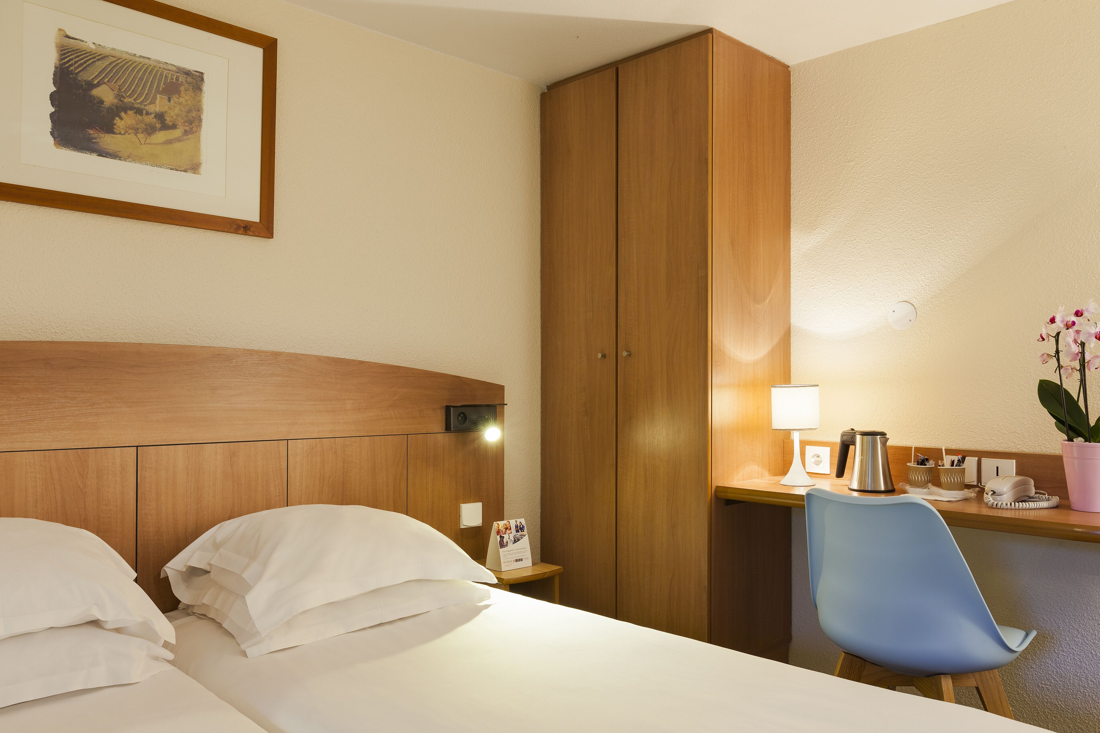 A hotel room with a bed, a desk with a chair, a wooden wardrobe, a framed picture on the wall, and a nightstand with a lamp and a phone.