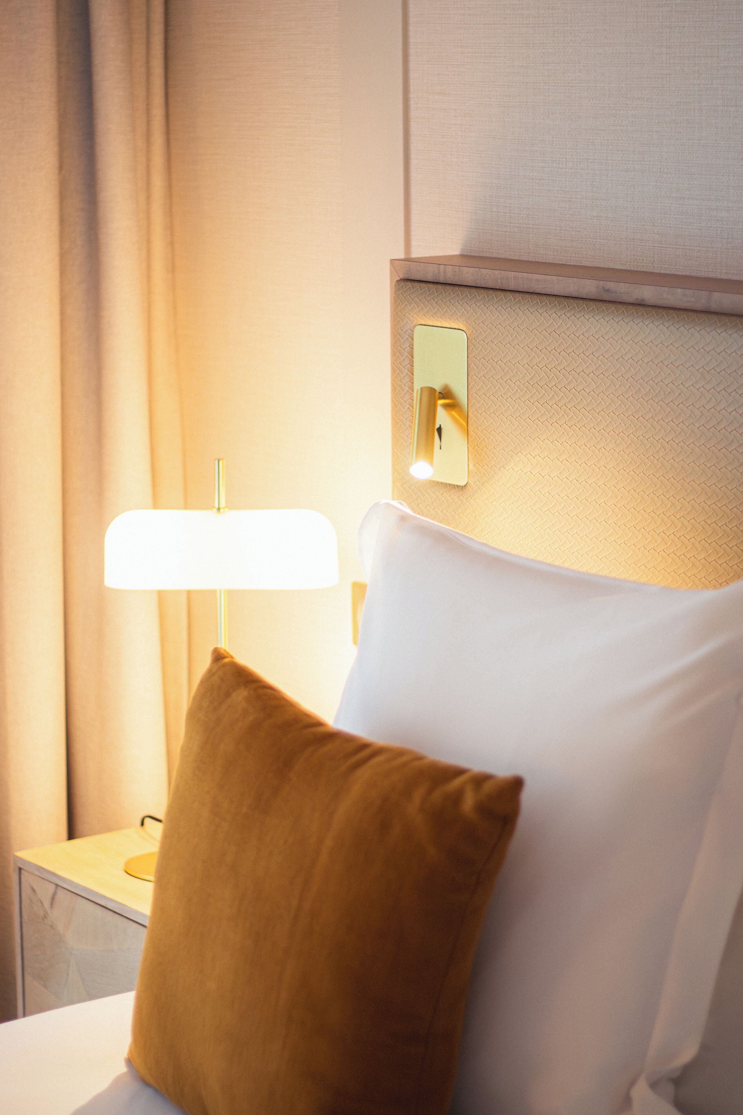 A hotel room with a bed, a nightstand, a lamp, and a wall-mounted light switch.